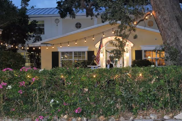 Evening view of a historic retirement residence with a yellow exterior, white trim, and black shutters. The building is partially obscured by a hedge with pink flowers. String lights are hung above the hedge, and an American flag is displayed near the entrance. A person is standing near the doorway under a lit archway.