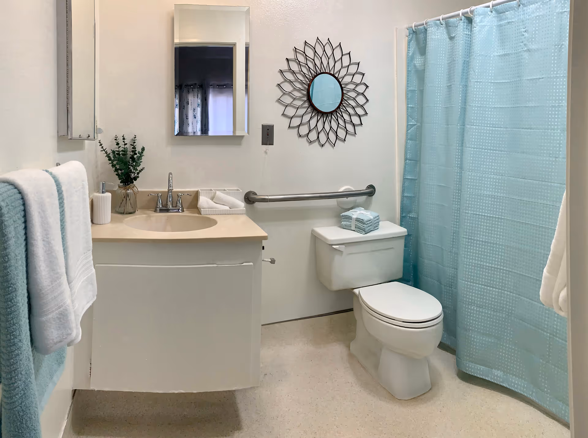 A clean and well-lit bathroom with a white toilet, a beige countertop with a sink, a wall-mounted mirror, a decorative round mirror with a petal-like frame, a grab bar next to the toilet, and a blue shower curtain. Towels in white and light blue are neatly hung and folded, and a small vase with green foliage is placed on the countertop.
