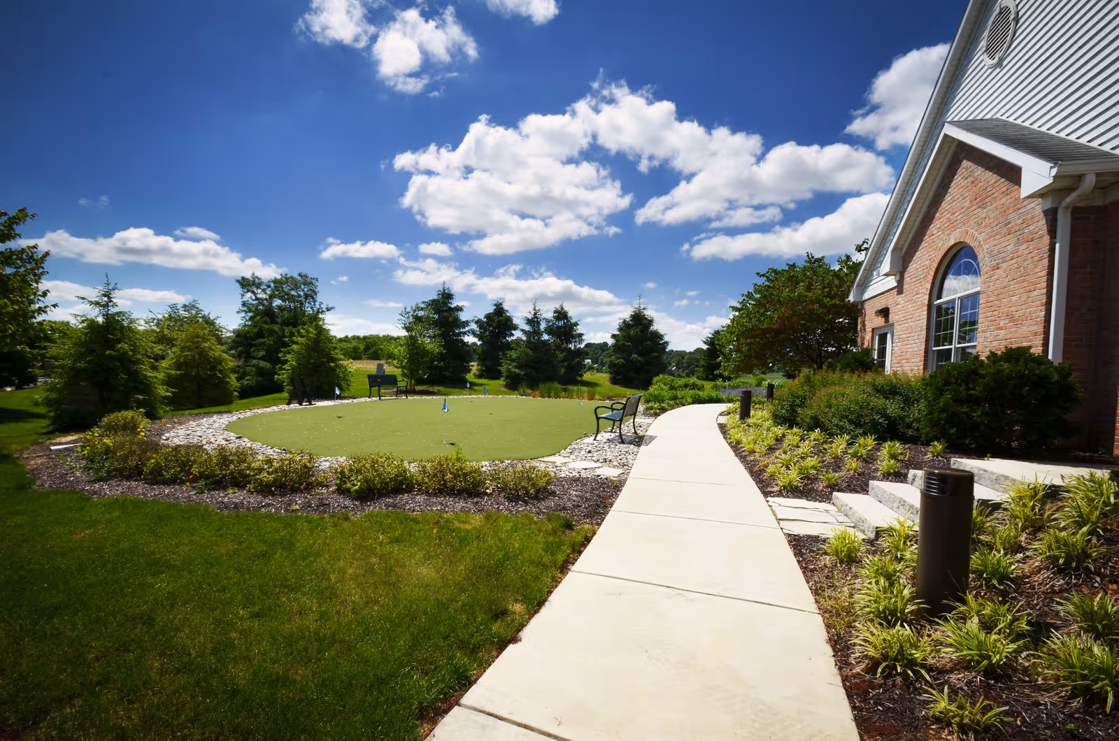 A bright outdoor scene at Luther Acres featuring a curved concrete walkway leading past a landscaped garden area with shrubs and small plants. To the right is a brick building with a large arched window. On the left side of the walkway is a circular putting green surrounded by rocks and greenery, with benches and trees in the background under a blue sky with scattered clouds.