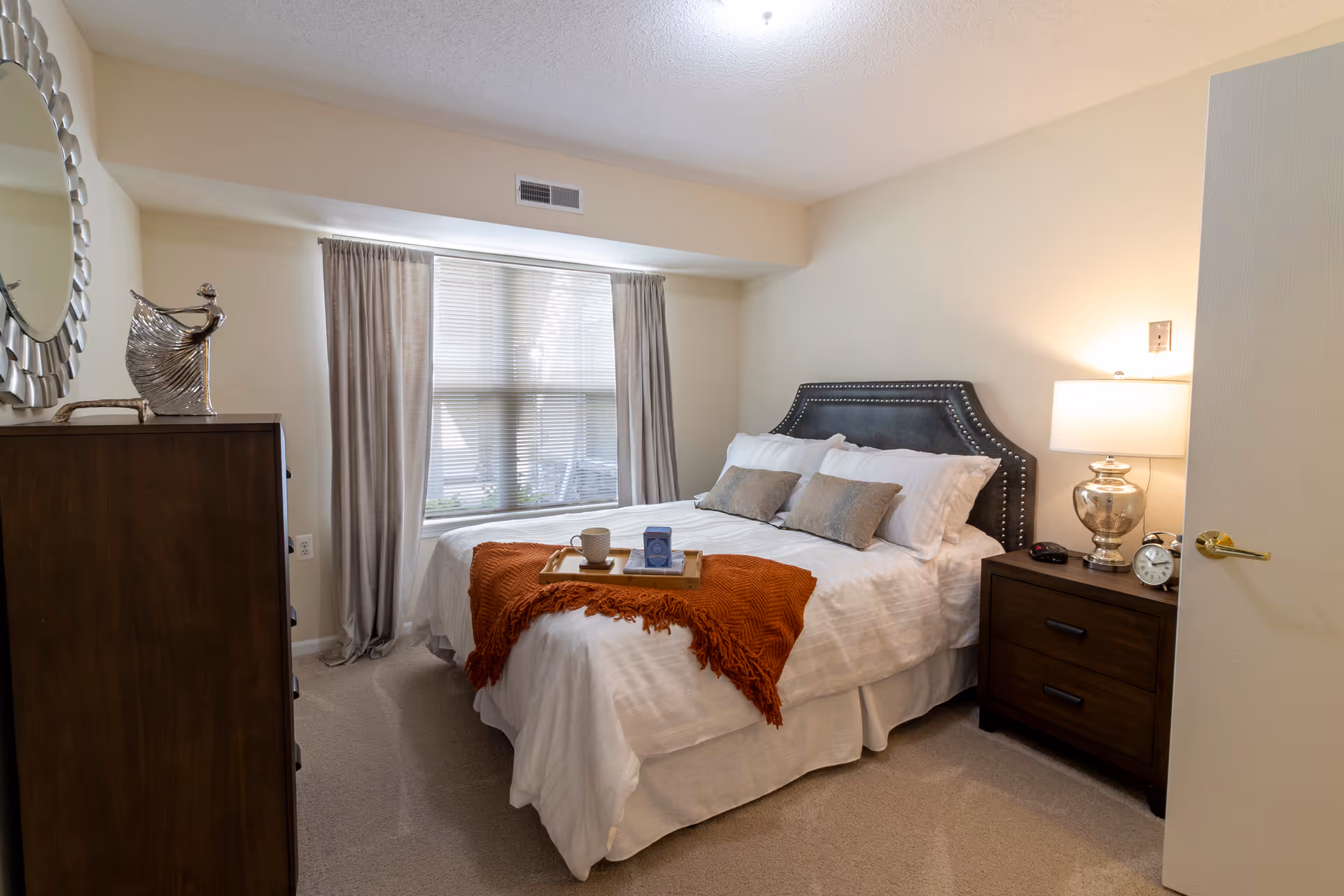 A cozy bedroom with a neatly made bed featuring white linens, two beige pillows, and an orange throw blanket. A wooden nightstand with a silver lamp, alarm clock, and remote control is beside the bed. A wooden dresser with a decorative silver sculpture and a round mirror hangs on the wall. A large window with beige curtains allows natural light into the room.