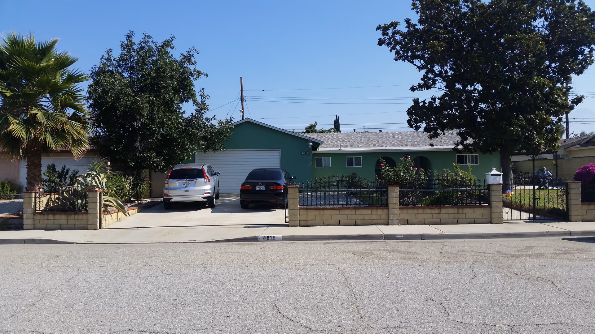 Front exterior view of a single-story green house with a gray roof, two cars parked in the driveway, a fenced front yard with trees and plants, and a clear blue sky.