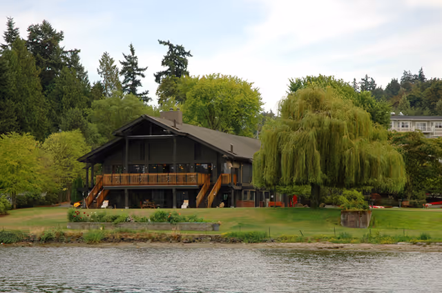 A large two-story building with a wooden balcony and stairs on both sides, situated by the water with a grassy lawn and a large weeping willow tree in front. The building is surrounded by various green trees and shrubs under a partly cloudy sky.