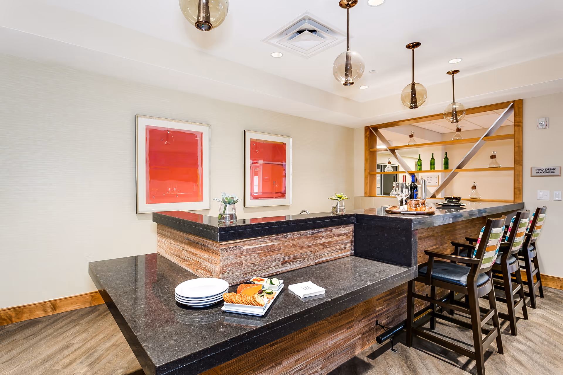 A modern indoor bar area with a black granite countertop and wooden paneling. There are four high chairs with colorful striped cushions lined up along the bar. On the counter, there are plates, a tray with crackers, cheese, and cucumber slices, wine glasses, and bottles of wine. The wall behind the bar has two framed abstract red artworks and a shelving unit with green bottles and decanters. The ceiling has recessed lighting and three pendant lights with round glass shades.