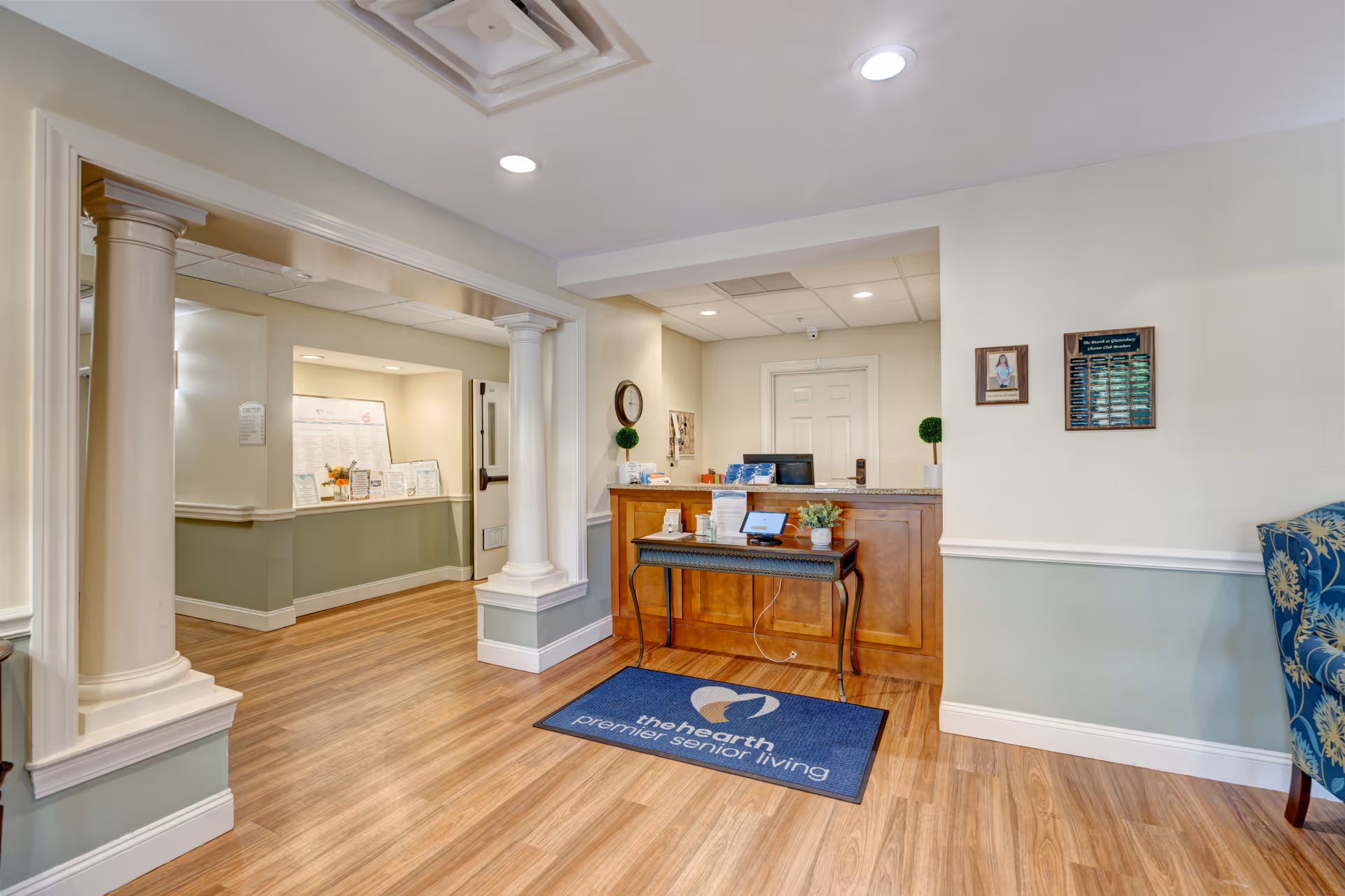 Reception desk and lobby area of a senior living facility with columns, wood flooring, and a welcome mat.