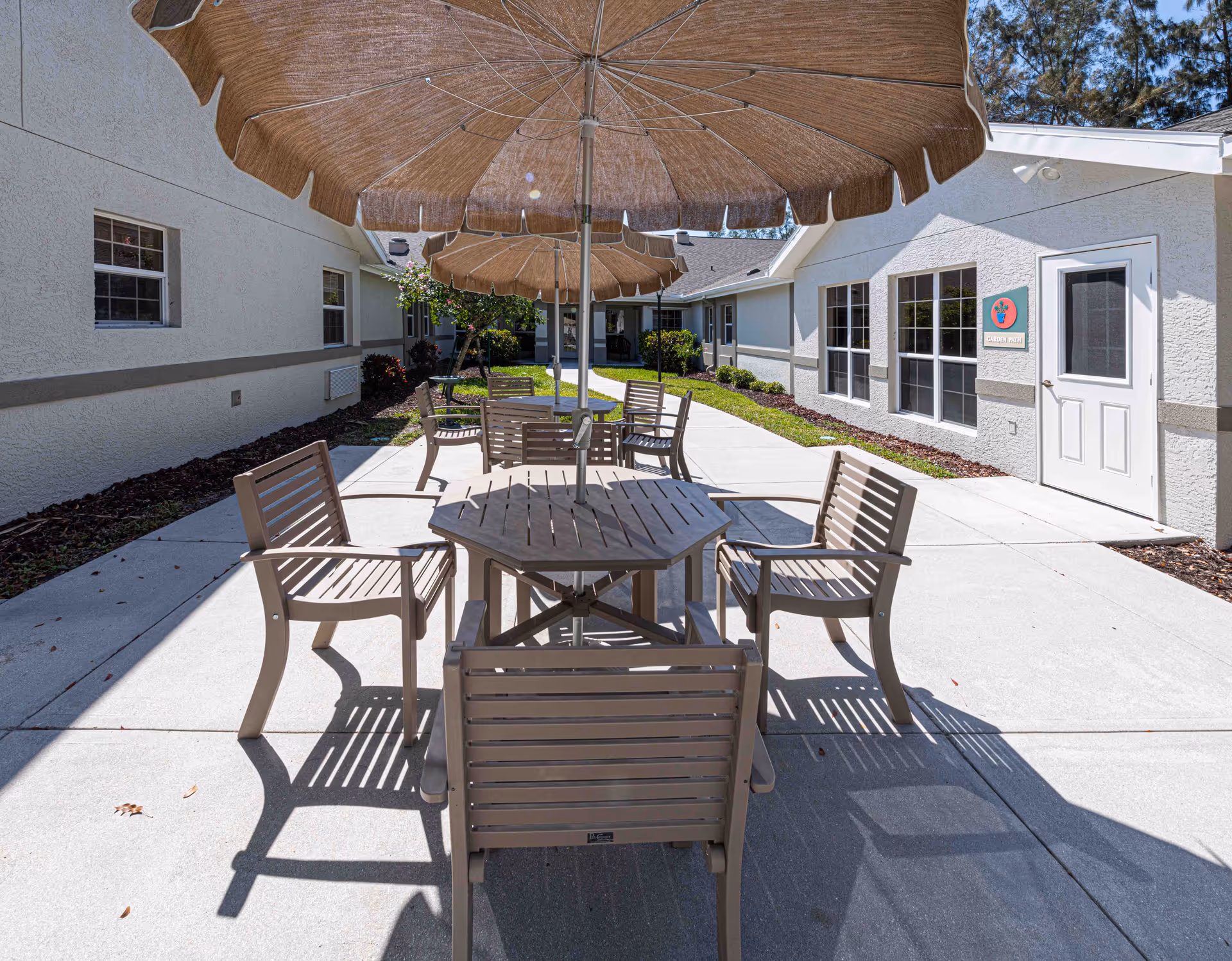 Outdoor patio area at Arden Courts - ProMedica Memory Care Community (Ft. Myers) featuring multiple wooden tables with matching chairs and large beige umbrellas providing shade. The patio is surrounded by the building with windows and a door visible, along with some landscaping including bushes and trees.