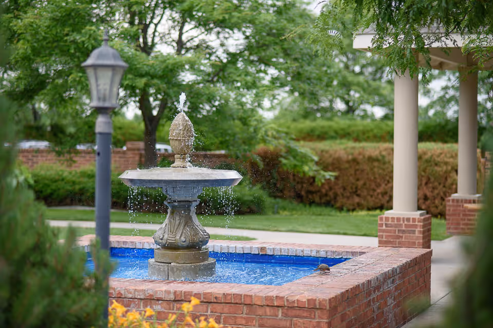 Outdoor garden area with a decorative stone water fountain in the center, surrounded by a brick border. There is a lamp post on the left side and green trees and bushes in the background.