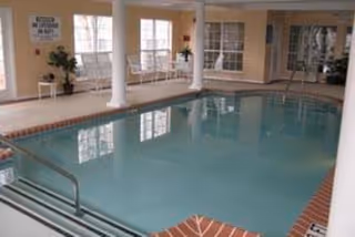 Indoor swimming pool area with clear water, surrounded by white columns and large windows letting in natural light. There are a few chairs and potted plants along the walls, and a metal handrail leading into the pool.