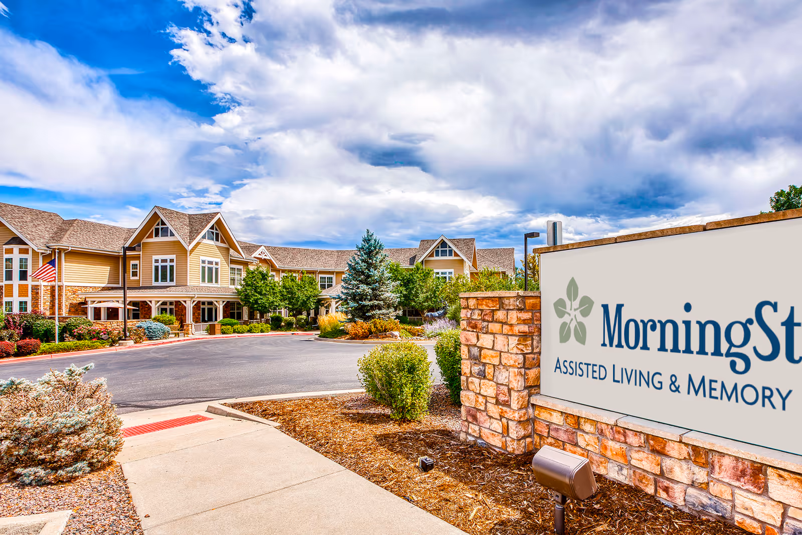 Exterior view of MorningStar Assisted Living & Memory Care at Applewood facility showing a large, multi-story building with beige siding and stone accents, surrounded by landscaped bushes and trees under a partly cloudy sky.