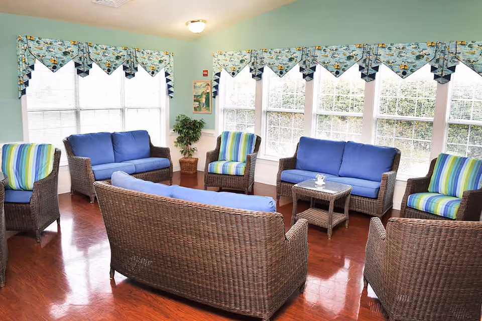A bright and cozy living room area with wicker furniture including two loveseats, three armchairs with blue and striped cushions, and a small wicker coffee table. The room has large windows with patterned valances, light blue walls, and polished wooden floors.
