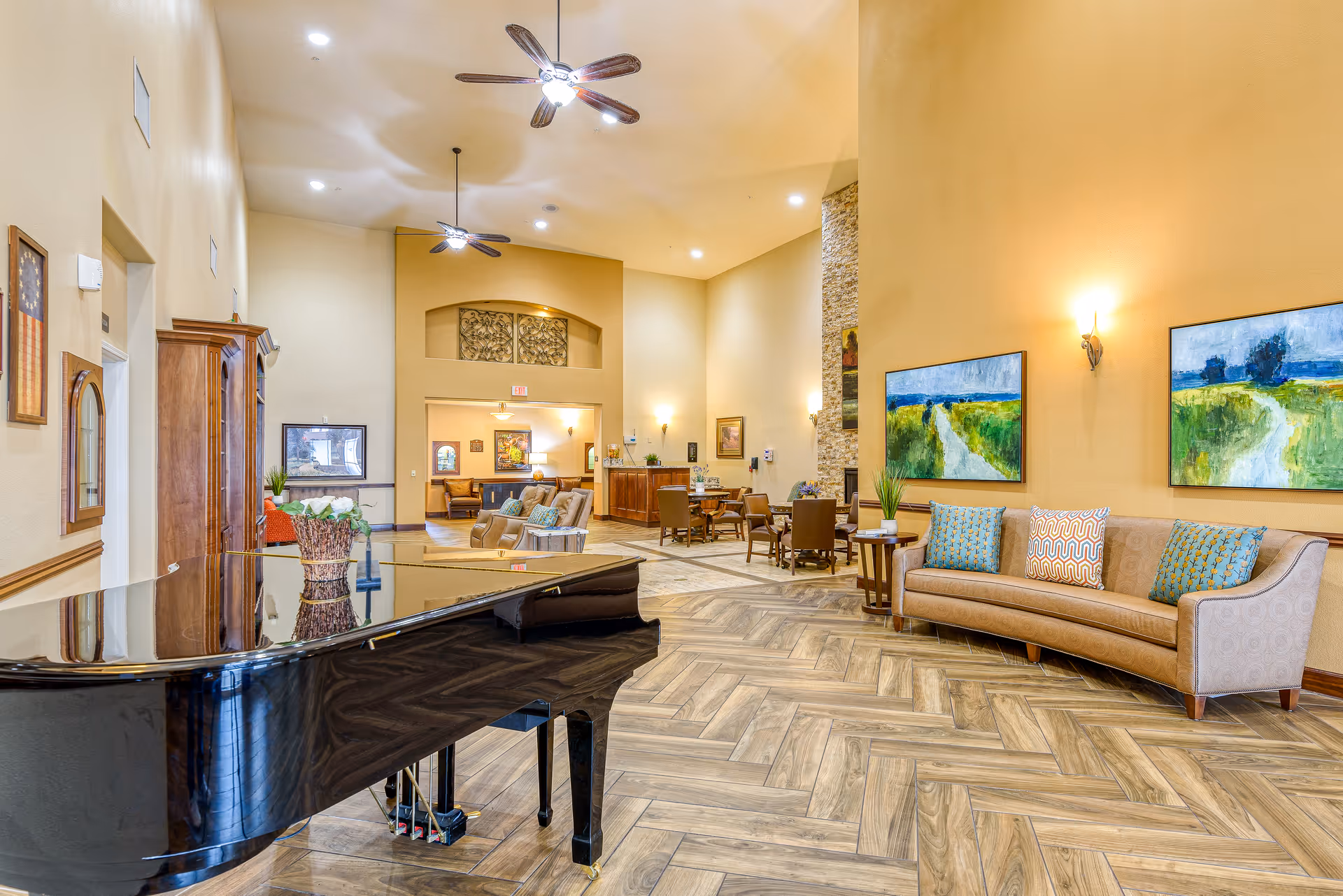 Spacious and well-lit common area in a senior living facility featuring a black grand piano in the foreground, a curved beige sofa with colorful cushions against the wall, two landscape paintings above the sofa, several tables and chairs in the background, ceiling fans, and warm yellow walls with wood-patterned flooring.