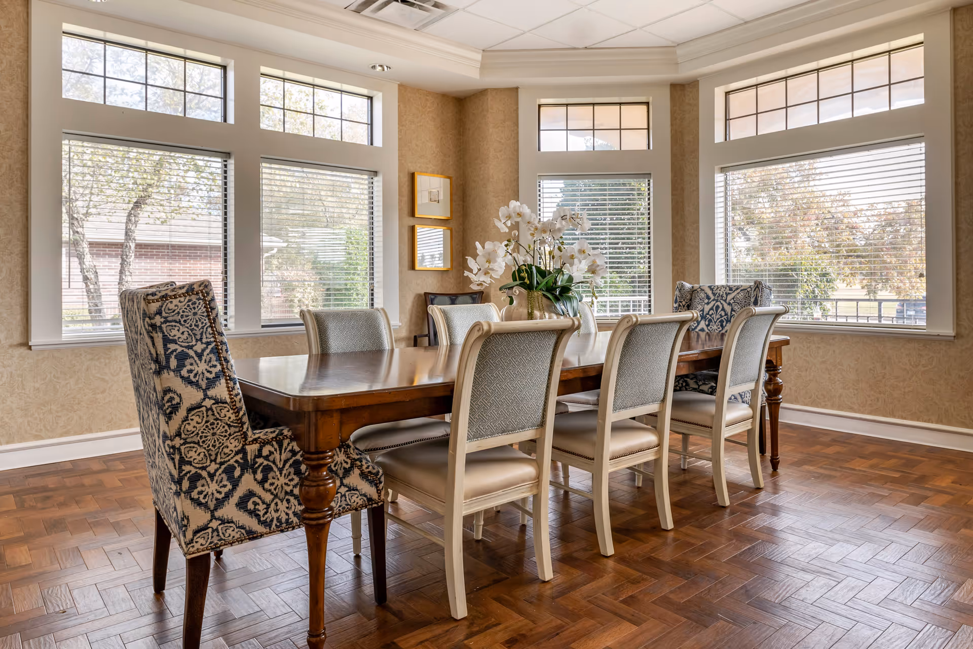 A bright dining room with a wooden dining table surrounded by eight chairs, including two patterned armchairs at the ends. The room has large windows with white blinds allowing natural light to fill the space, and a vase with white flowers is placed in the center of the table. The floor is wooden parquet, and the walls are beige with a subtle pattern.