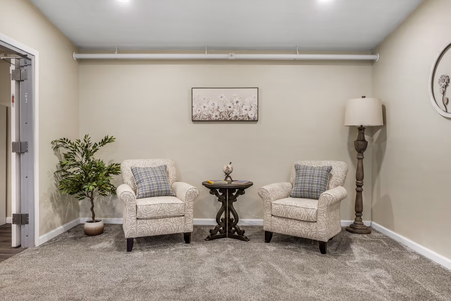 A cozy sitting area with two patterned armchairs each with a plaid cushion, a small round wooden table between them holding a small globe and a book, a potted plant on the left, a tall floor lamp on the right, and a framed floral artwork on the beige wall behind.