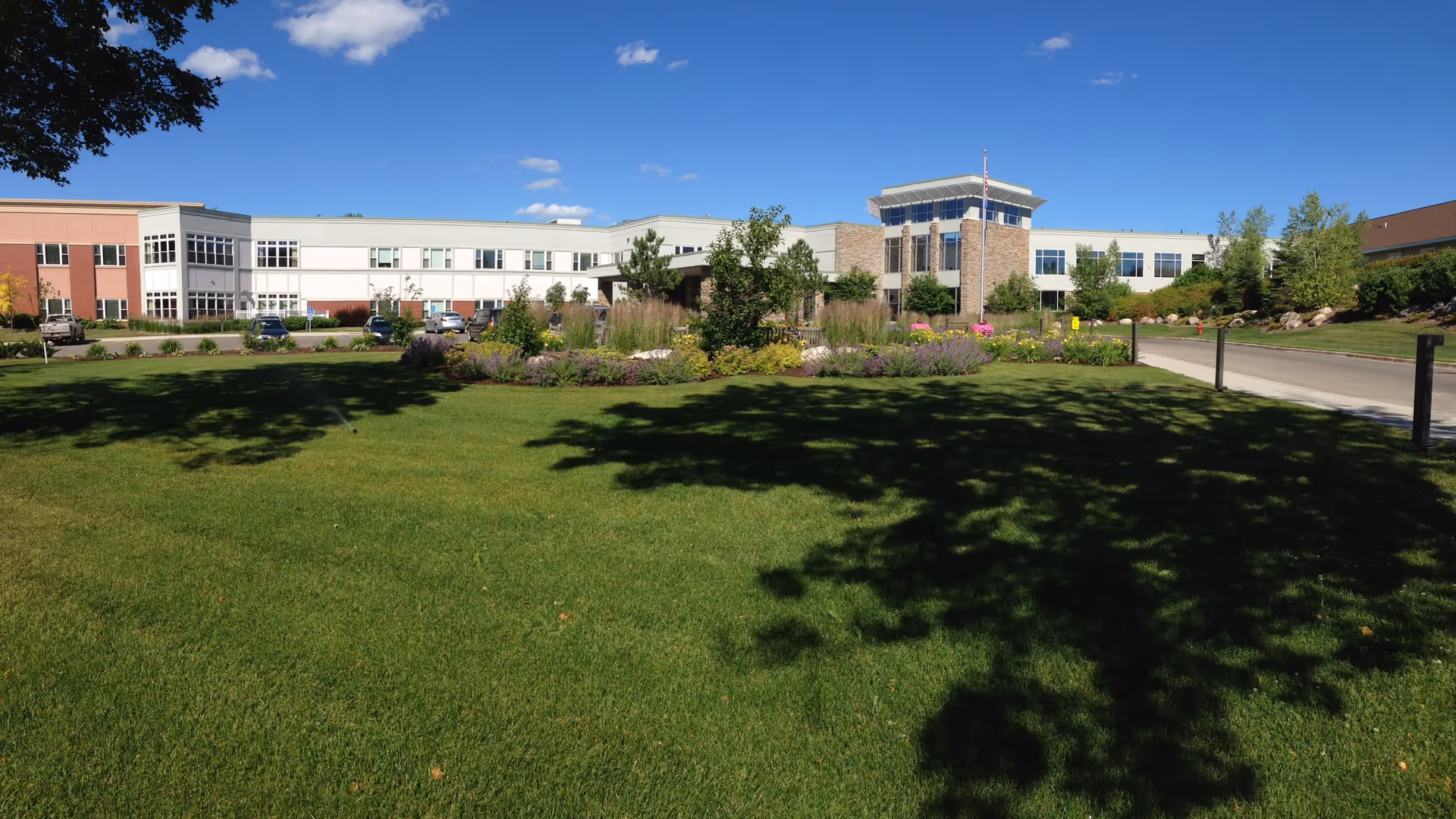 Wide exterior view of PioneerCare Center showing a large green lawn with trees and landscaped garden beds in front of a modern two-story building under a clear blue sky.