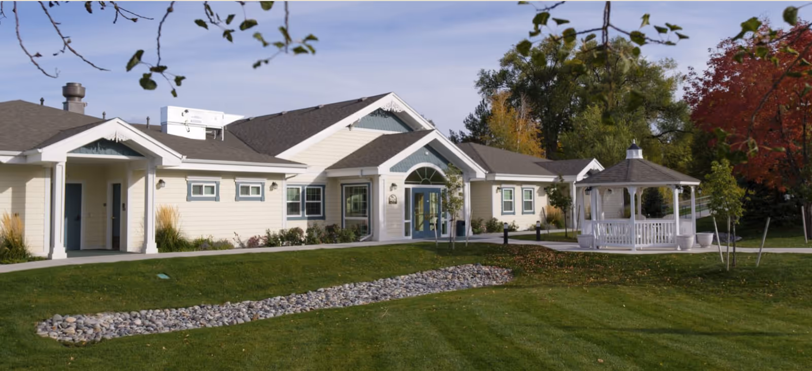 Exterior view of a single-story senior living facility building with cream-colored siding and blue trim. The building has multiple windows and a main entrance with double doors under a peaked roof. In front of the building is a well-maintained lawn with a dry rock creek bed and a white gazebo surrounded by small trees and shrubs. The sky is clear with some tree branches visible in the foreground.