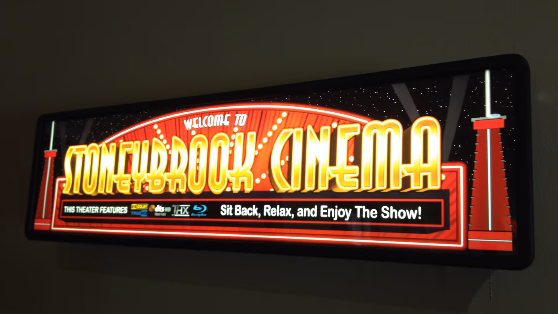 Illuminated sign reading 'Welcome to StoneyBrook Cinema' with a retro design, featuring the message 'Sit Back, Relax, and Enjoy The Show!' and icons indicating theater features such as Dolby TrueHD, dts HD, THX, and Blu-ray.