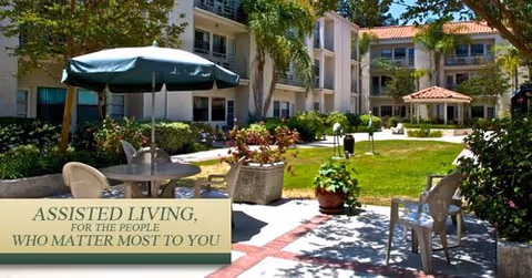Sunny courtyard with patio tables, chairs and an umbrella surrounded by landscaped lawns and apartment-style buildings.