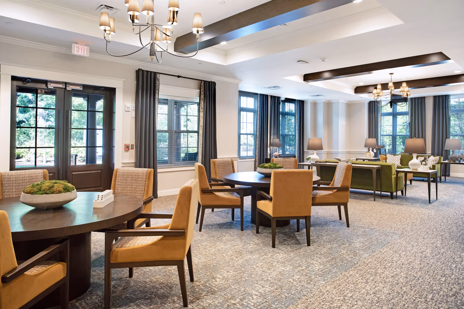 A spacious and well-lit common area in a senior living facility featuring round tables with cushioned chairs, large windows with dark curtains, green sofas, side tables with lamps, and elegant ceiling light fixtures. The room has a neutral color palette with beige walls and carpeted floors.