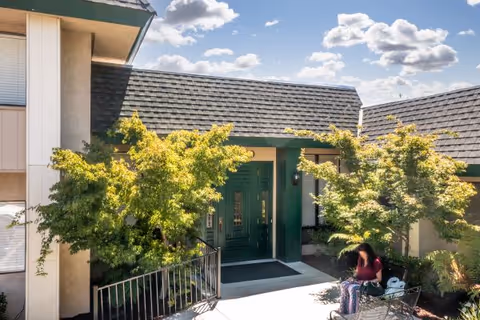 Entrance to a building with a green door and a shingled roof, surrounded by green trees and bushes. A person is sitting on a bench near the entrance under a sunny sky with scattered clouds.