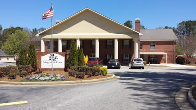 Front exterior view of Summer Village Magnolia Place building with a covered entrance, an American flag on a flagpole, several parked cars, and landscaped bushes and flowers in front.