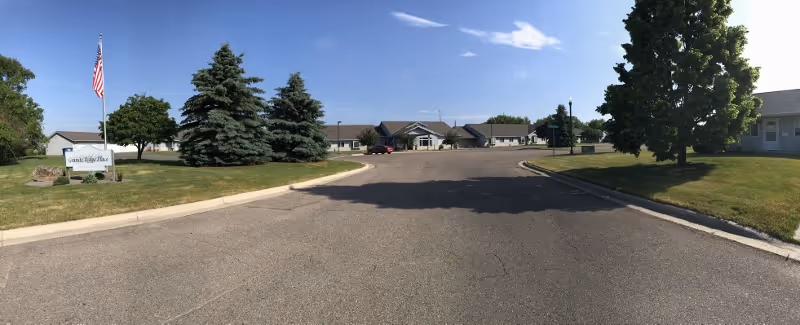 Wide view of the entrance driveway leading to a single-story senior living facility building with a manicured lawn, several trees, and an American flag on a flagpole near a sign that reads Granite Ridge Senior Living of Granite Falls Health under a clear blue sky.