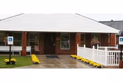 Entrance of a single-story brick building with a white roof and a covered porch. There are two handicap parking signs visible near the entrance, a white picket fence on the right side, and yellow flowers lining the walkway leading to the door.