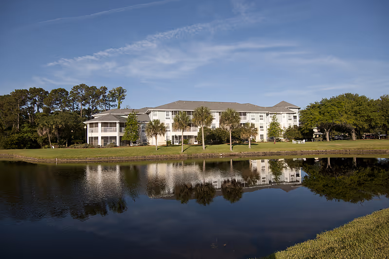 A large, white multi-story senior living facility building with a gray roof, surrounded by palm trees and other greenery, reflected in a calm pond in the foreground under a blue sky with light clouds.