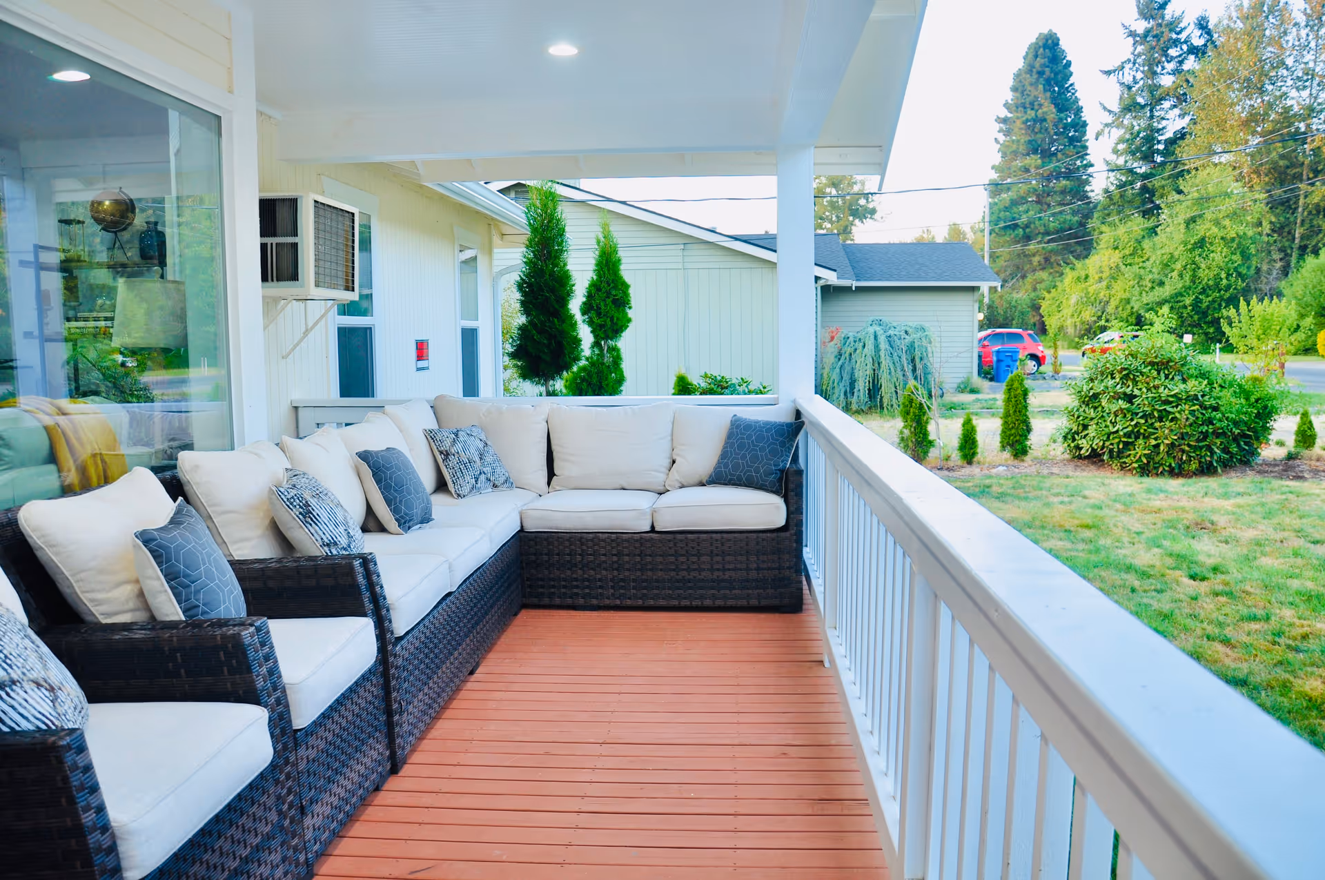Covered outdoor porch area with a large L-shaped wicker sofa with beige cushions and multiple decorative pillows. The porch has a wooden floor and white railing, overlooking a green yard with bushes and trees. A house wall with windows and an air conditioning unit is visible on the left side.