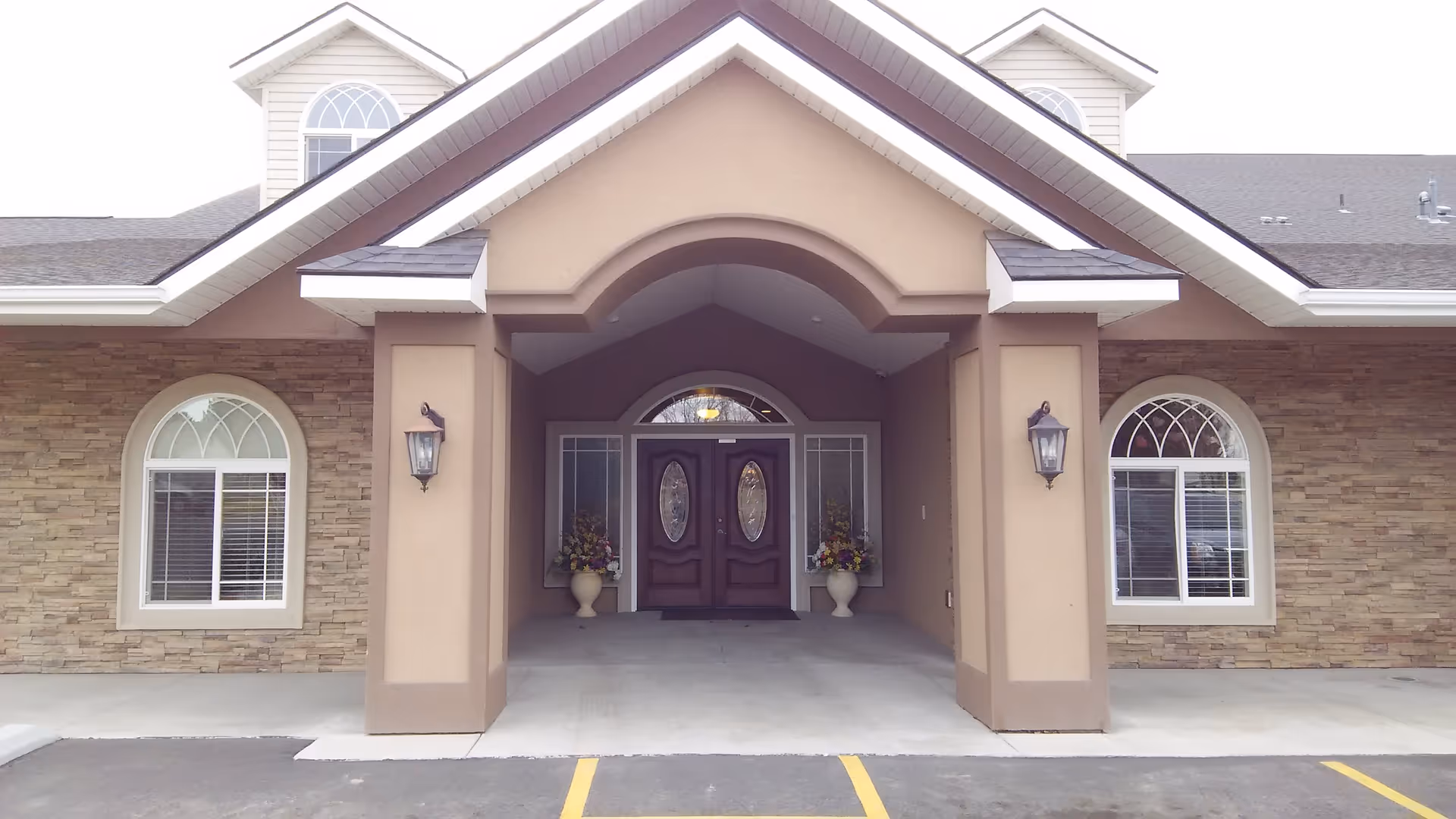 Front entrance of a building with a covered porch supported by two columns, featuring double wooden doors with oval glass panels, two large flower pots with colorful flowers on either side of the doors, and two arched windows with white trim and blinds on the stone and beige exterior walls.
