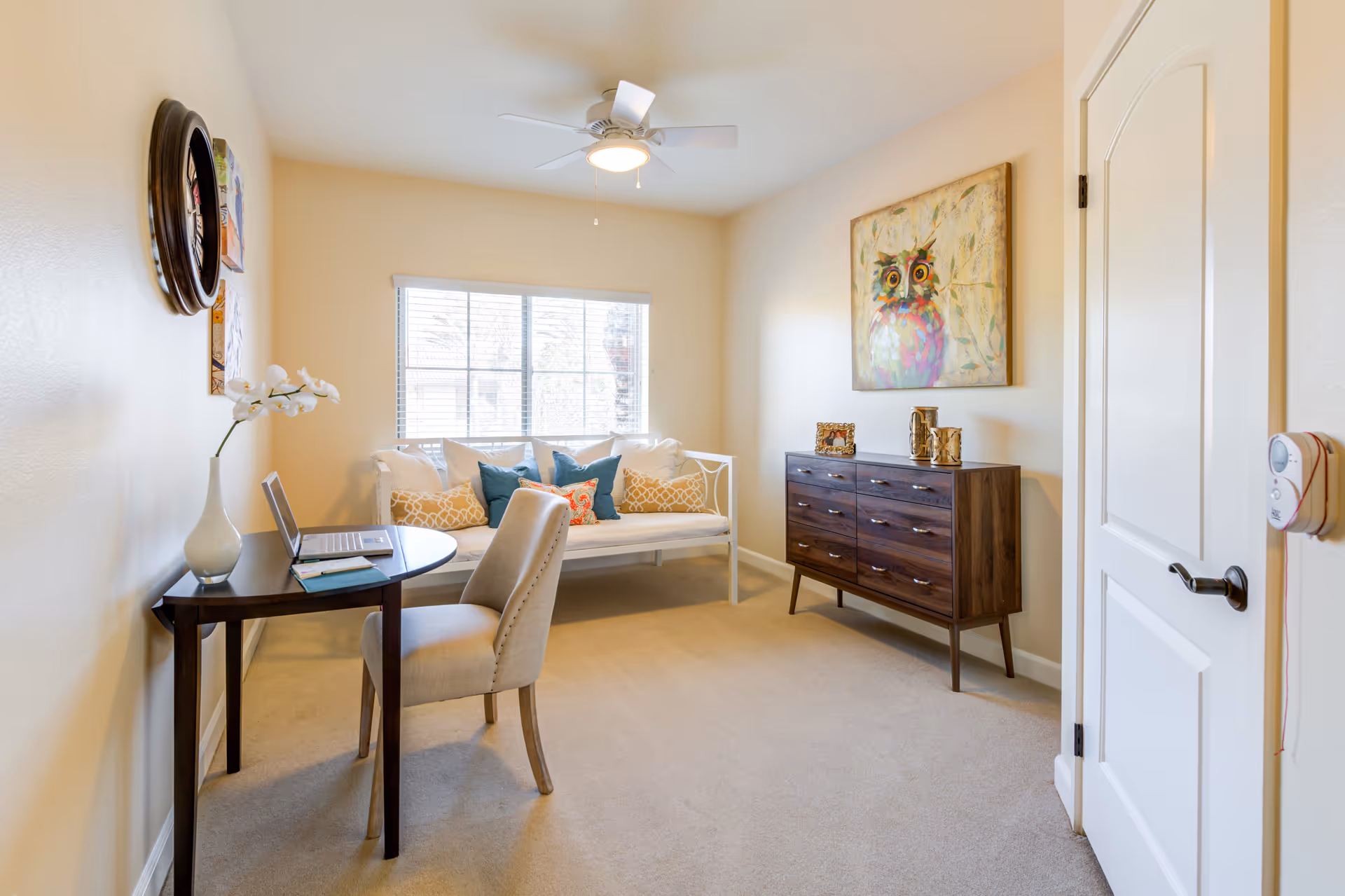 A bright room with beige walls and carpet featuring a small dark wood desk with a laptop and a vase with white flowers, a beige upholstered chair, a white cushioned bench with multiple decorative pillows, a dark wood dresser with decorative items, a colorful owl painting on the wall, and a ceiling fan with light. A window with blinds lets in natural light.