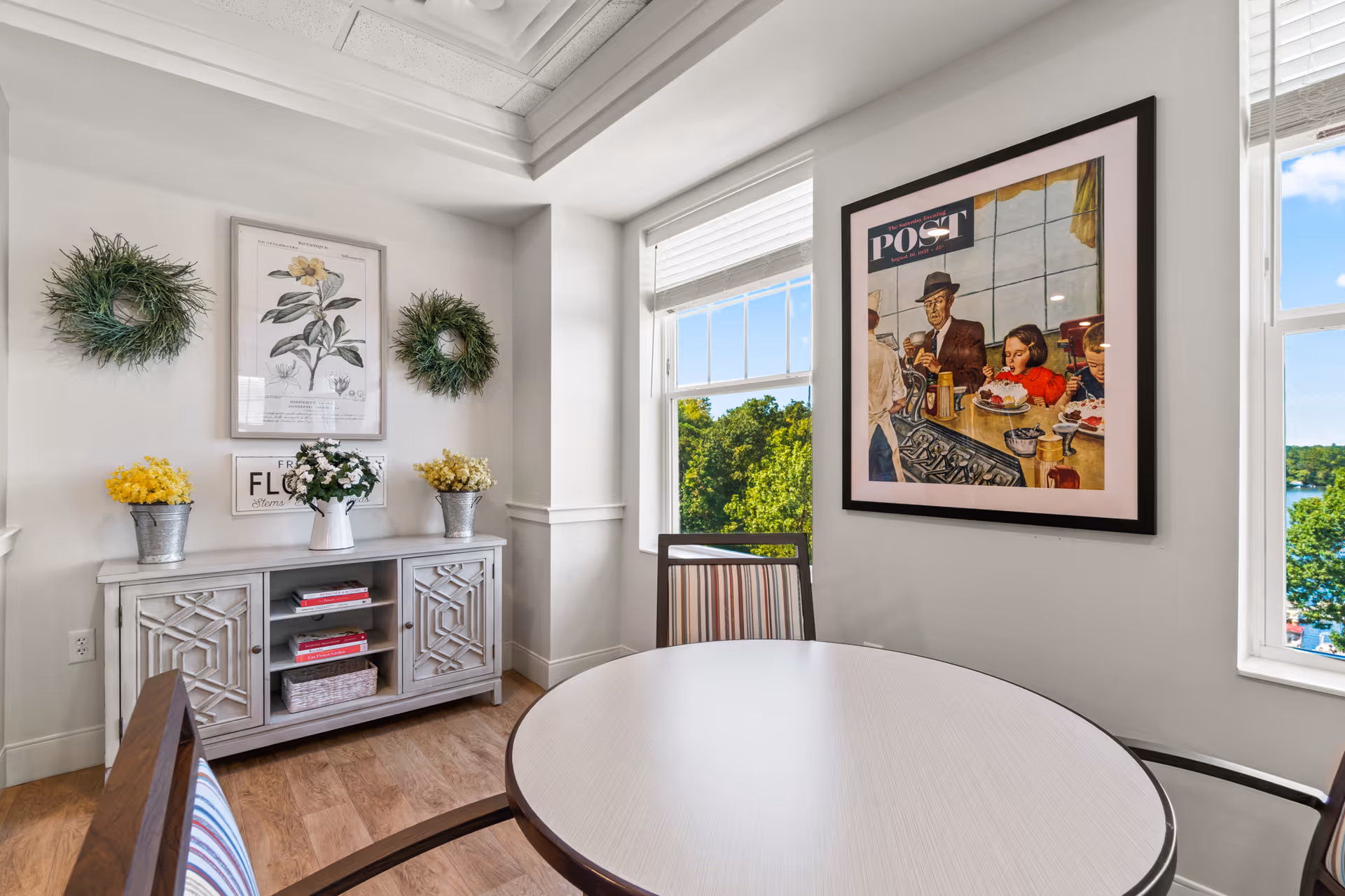 A bright dining area with a round table and striped chairs. The room features two windows with views of green trees and a body of water. On the wall is a framed vintage-style poster of a man and children at a diner counter. Opposite the table is a decorative cabinet with floral arrangements and two green wreaths hanging on the wall.