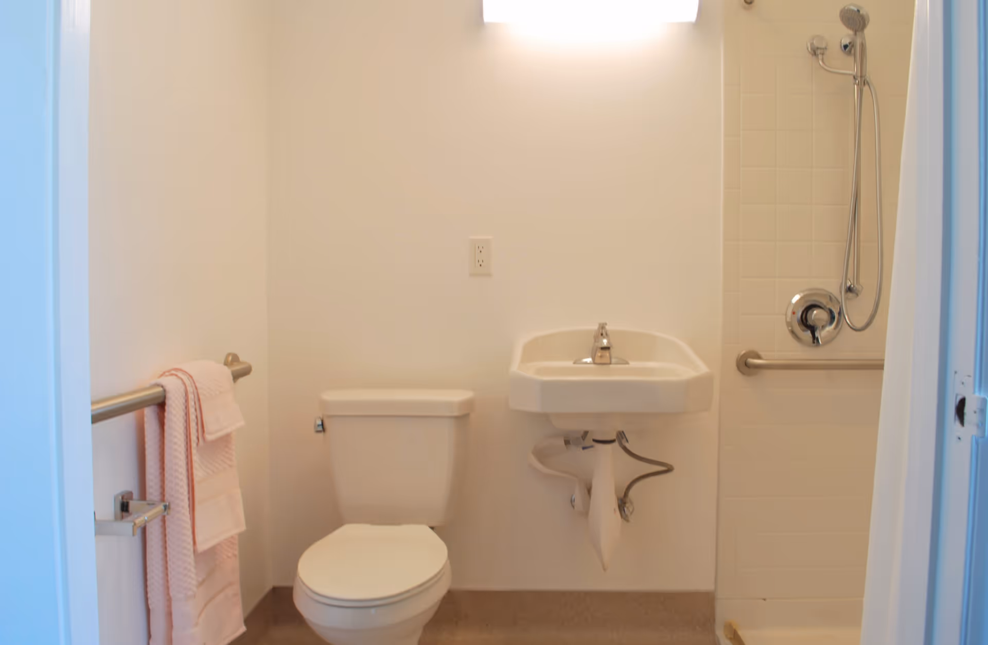 A clean and simple bathroom featuring a white toilet, a wall-mounted white sink with exposed plumbing, a shower area with a handheld showerhead and grab bar, and a towel rack with pink towels on the left wall.