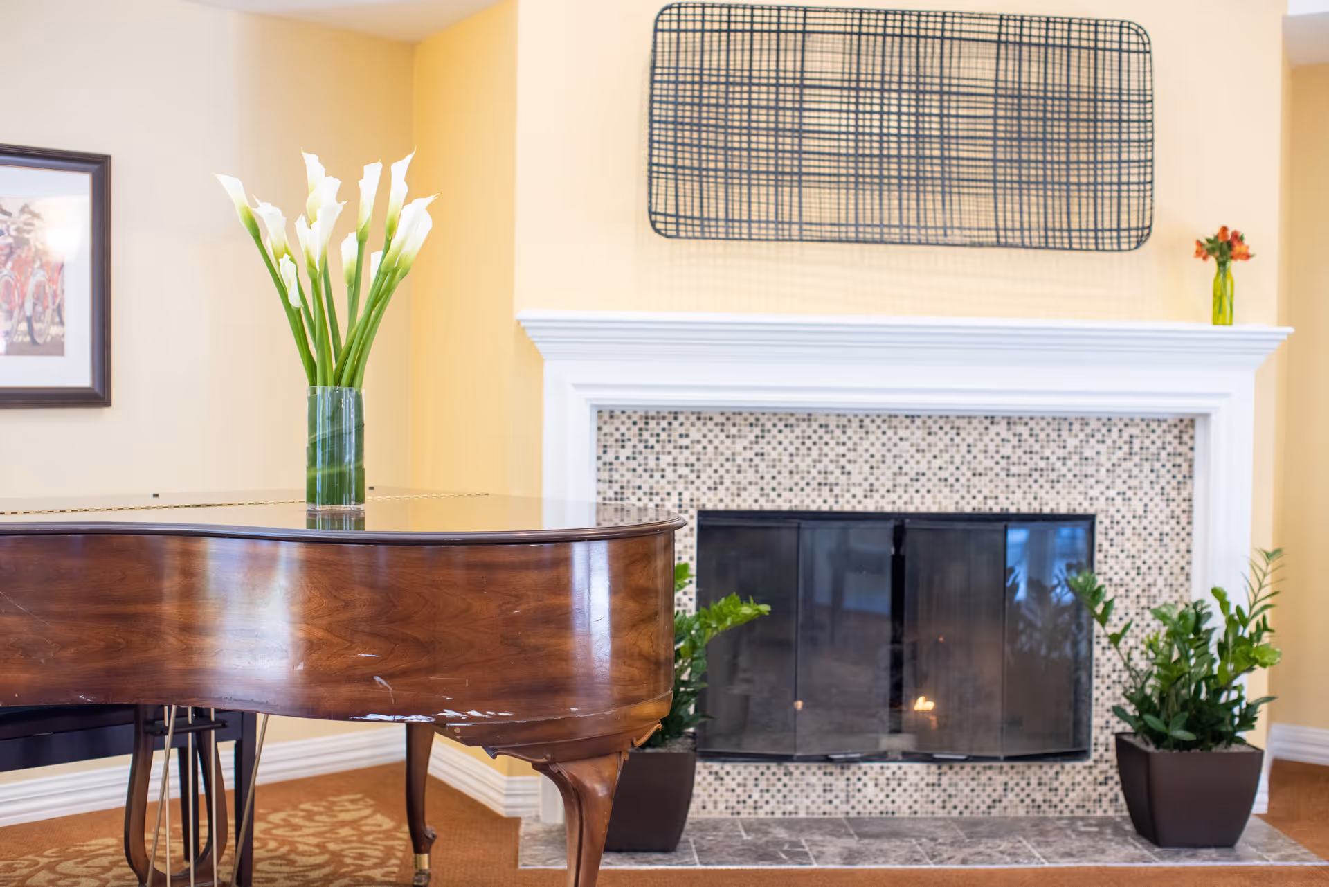 A cozy interior space featuring a polished wooden grand piano with a vase of white calla lilies on top, positioned near a fireplace with a mosaic tile surround and a white mantel. Two potted green plants flank the fireplace, and a modern rectangular wall art piece hangs above it. The walls are painted a soft yellow, and a framed picture is partially visible on the left.