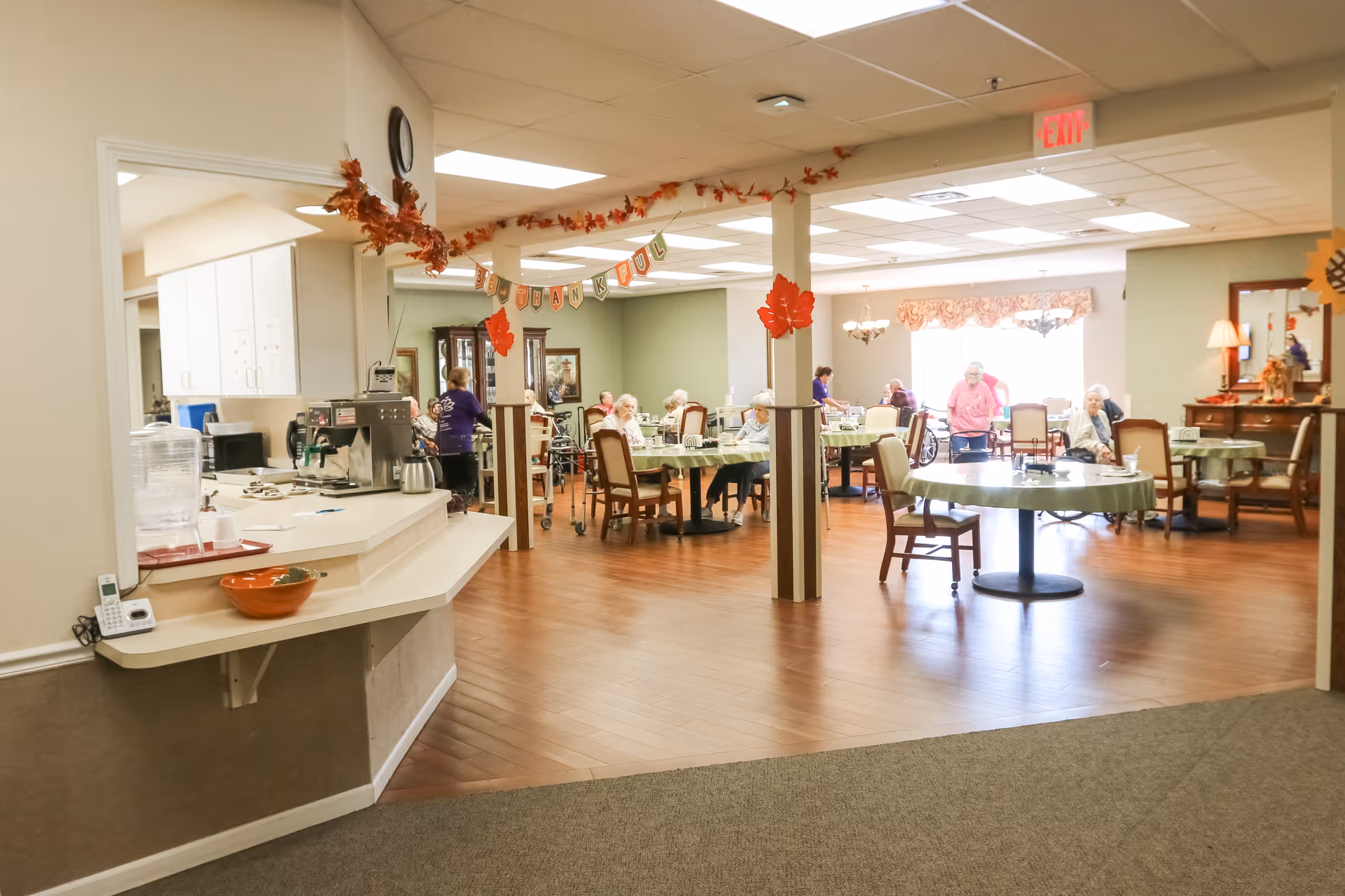 A dining room in a senior living facility decorated with autumn leaves and a banner that says 'BE THANKFUL'. Several elderly residents are seated at round tables covered with green tablecloths, and a few staff members are assisting them. The room has wooden flooring, soft lighting, and a cozy atmosphere.