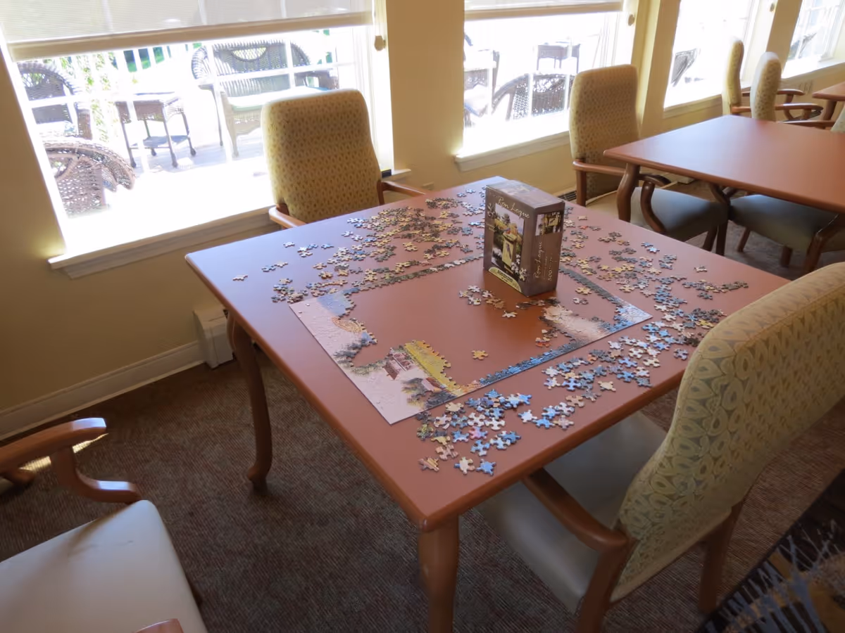 Sunlit activity room table with an unfinished jigsaw puzzle and several chairs by large windows.