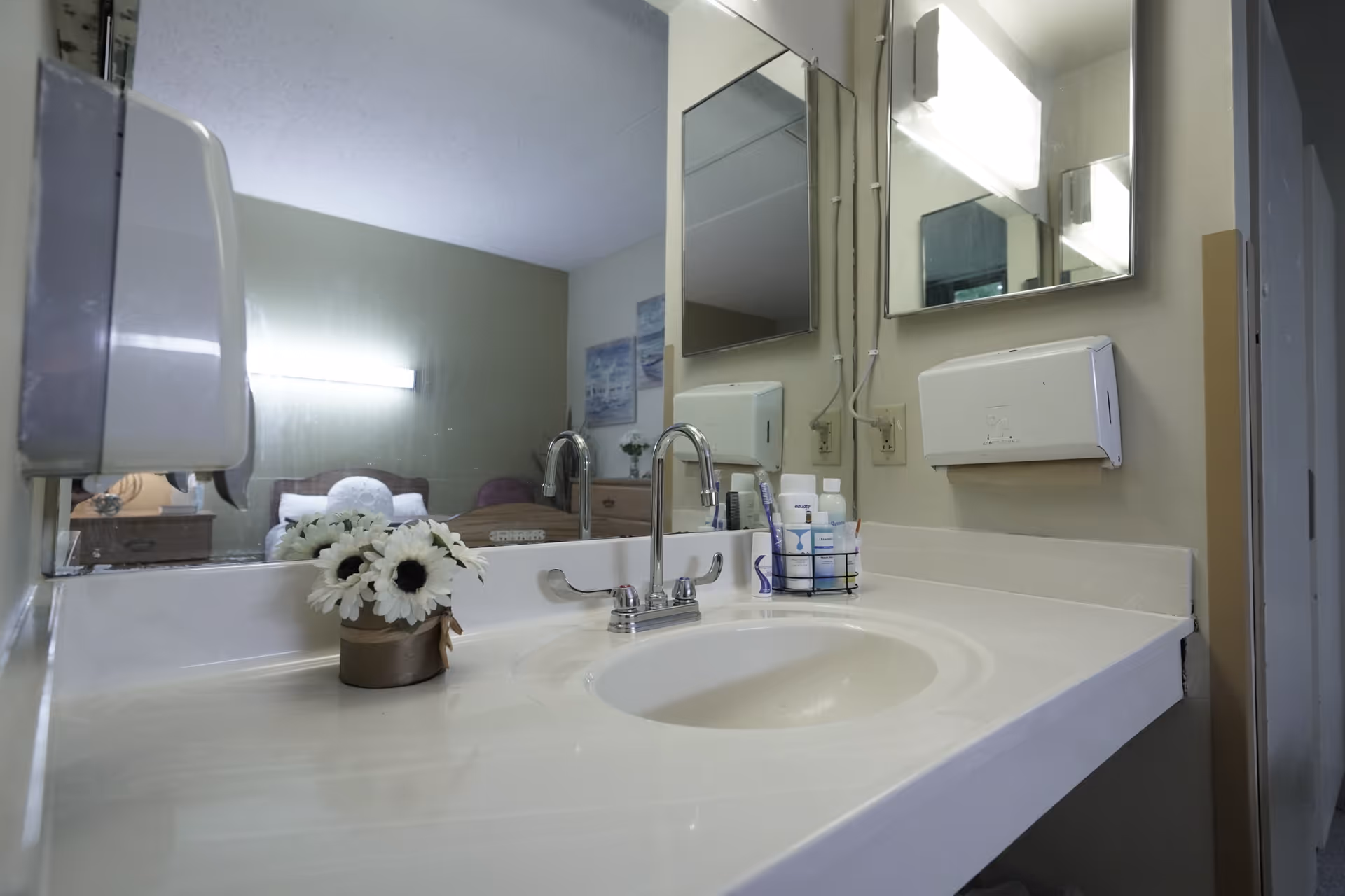 A clean bathroom sink area with a white countertop, a silver faucet, a small vase with white flowers, and toiletries in a holder. A large mirror reflects a bedroom with a bed, nightstand, and wall art. The bathroom has a wall-mounted soap dispenser and paper towel dispenser.