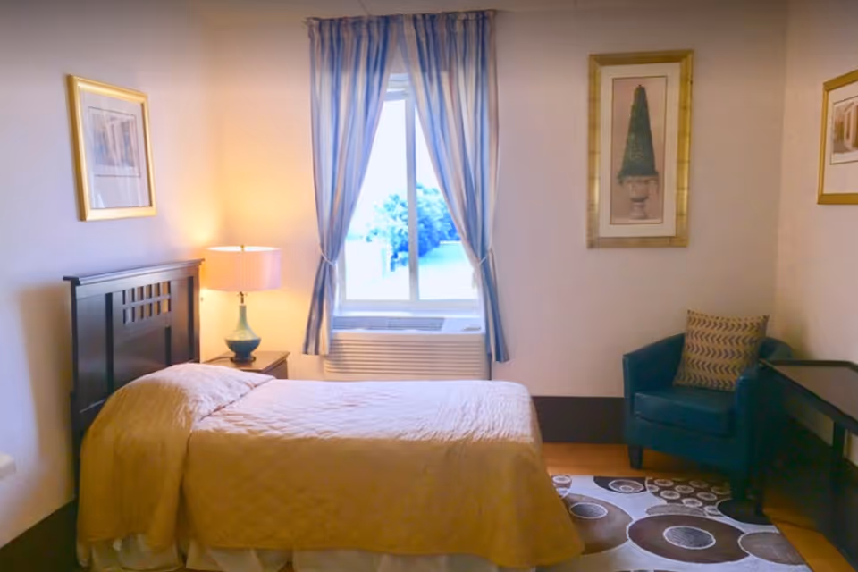 A bedroom with a single bed covered in a beige quilt, a bedside table with a lamp, a window with blue and white striped curtains, a blue armchair with a patterned cushion, framed artwork on the walls, and a patterned rug on the floor.