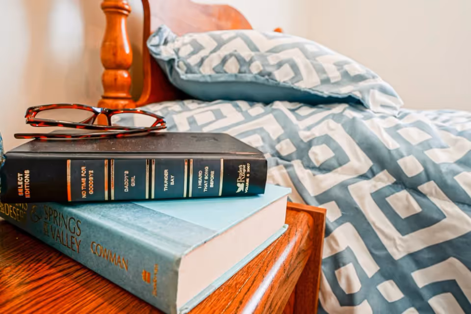 Close-up of a wooden bedside table with two stacked books and a pair of tortoiseshell reading glasses on top. In the background, a bed with a wooden headboard and blue and white geometric patterned bedding is visible.