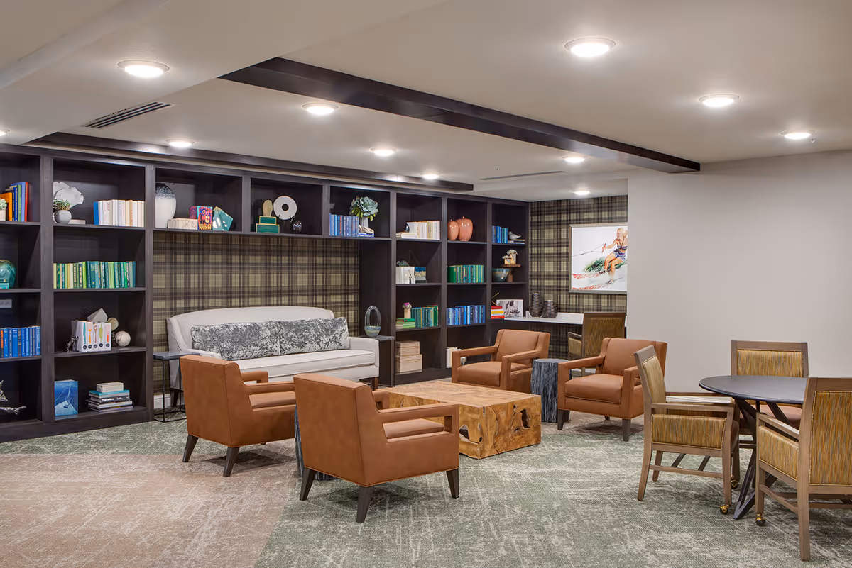 A cozy living room area in a senior living facility with a beige sofa, four brown armchairs, a wooden coffee table, and a round table with chairs. The back wall features dark wood built-in shelves filled with books and decorative items, and the wall behind the sofa has a plaid pattern. The room is well-lit with ceiling lights.