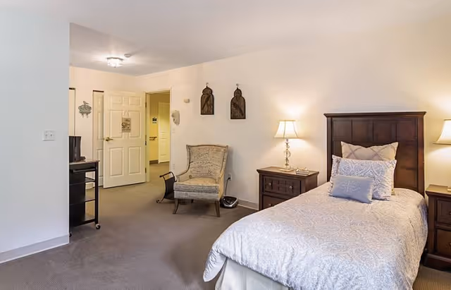 A senior living facility bedroom with a single bed dressed in light-colored bedding, two wooden nightstands with lamps on either side, a cushioned armchair, and a TV on a stand near the open door leading to a hallway.