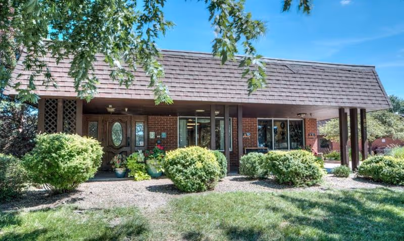 Exterior view of Rossville Healthcare and Rehabilitation Center showing a brick building with a brown shingled roof, large windows, and a covered entrance. The foreground features green bushes, plants, and a grassy area under a clear blue sky.