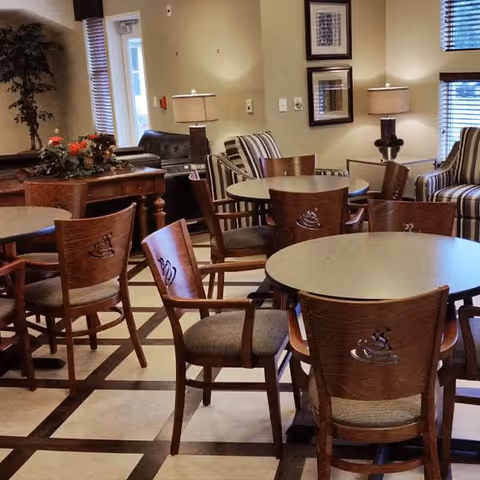 A cozy common area in a senior living facility featuring round tables with wooden chairs arranged around them. The chairs have a decorative design on the backrests. In the background, there are striped armchairs, table lamps, framed artwork on the walls, and a wooden side table with a floral arrangement. Large windows with blinds allow natural light into the room.