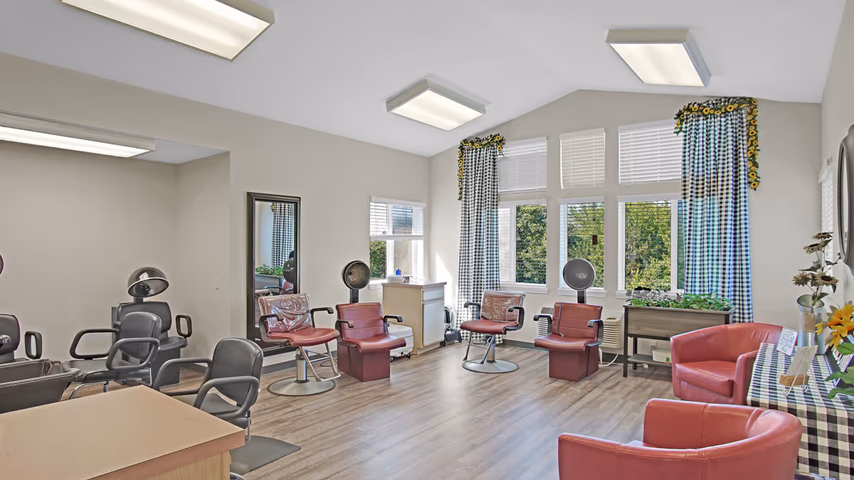 Interior of a hair salon area in a senior living facility with several salon chairs, hair dryers, large windows with checkered curtains, and a couple of red armchairs near a table with a checkered tablecloth and flowers.