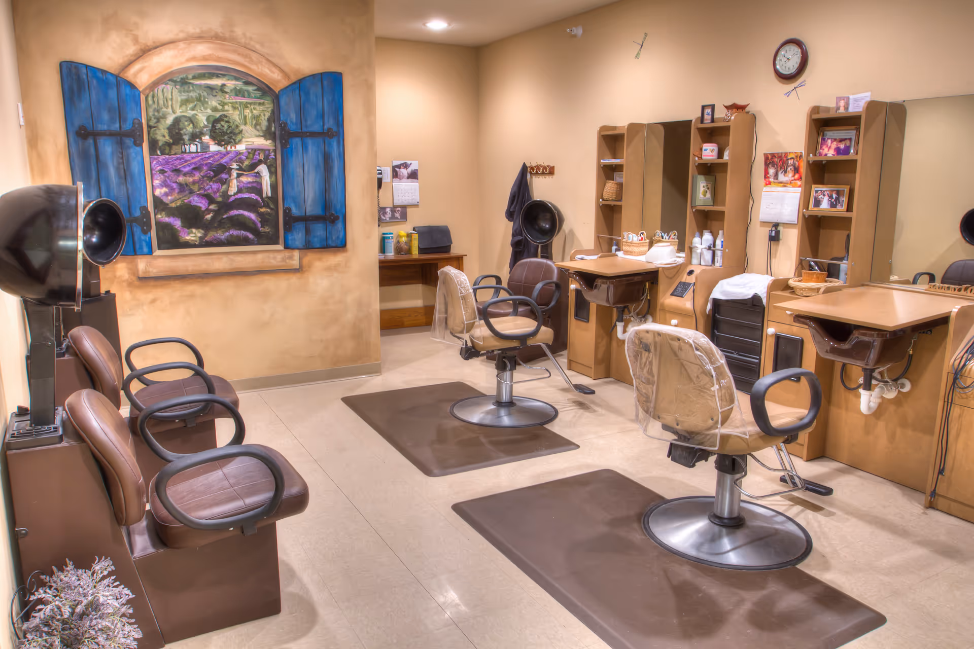 Interior salon area with multiple styling chairs, hair dryers, workstations, and a decorative mural painting of a window looking onto lavender fields.