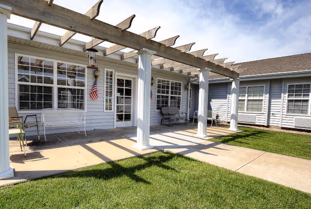 Outdoor patio area with a pergola, benches, and a manicured lawn in front of a light-gray building.