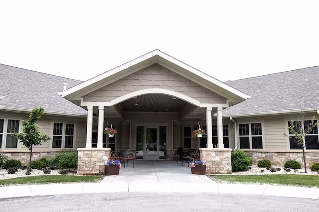 Front entrance of a single-story assisted living facility with a covered porch supported by white columns. The building has beige siding with stone accents at the base and multiple windows. There are hanging flower baskets and potted plants near the entrance, with a paved driveway and small landscaped areas with grass and shrubs.