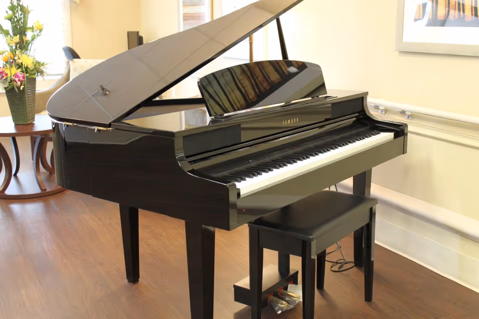 A black grand piano with a bench in a communal living area with wood floors and a flower arrangement in the background.