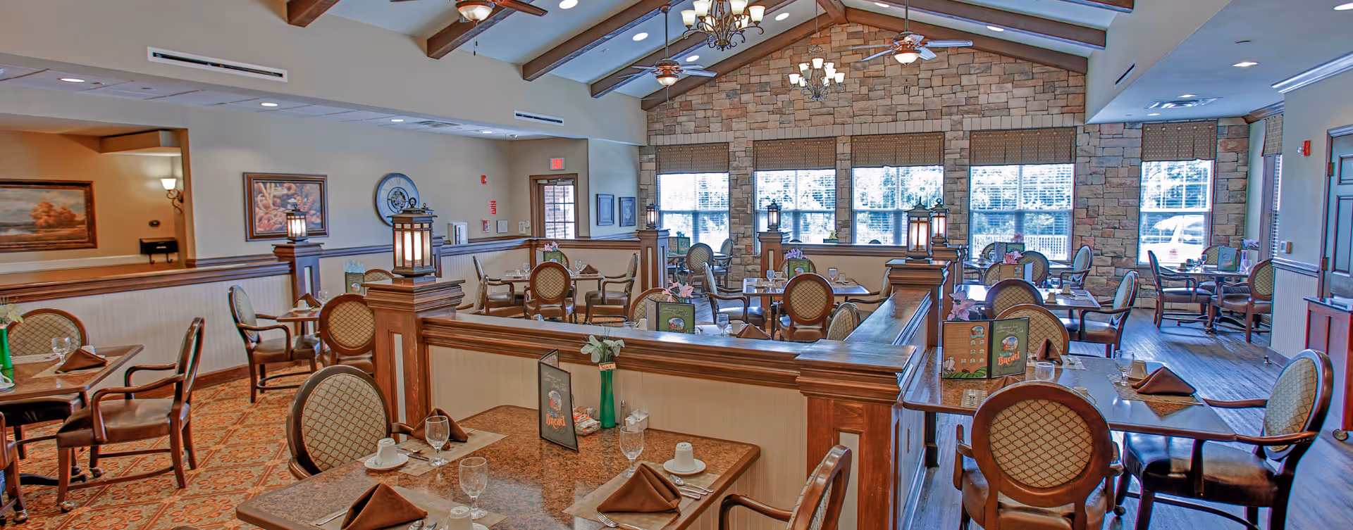 A spacious dining room with multiple tables set with brown napkins, glasses, and menus. The room features wooden chairs with cushioned seats and backs, a stone accent wall with large windows letting in natural light, ceiling fans, and chandeliers hanging from a vaulted ceiling with exposed wooden beams.