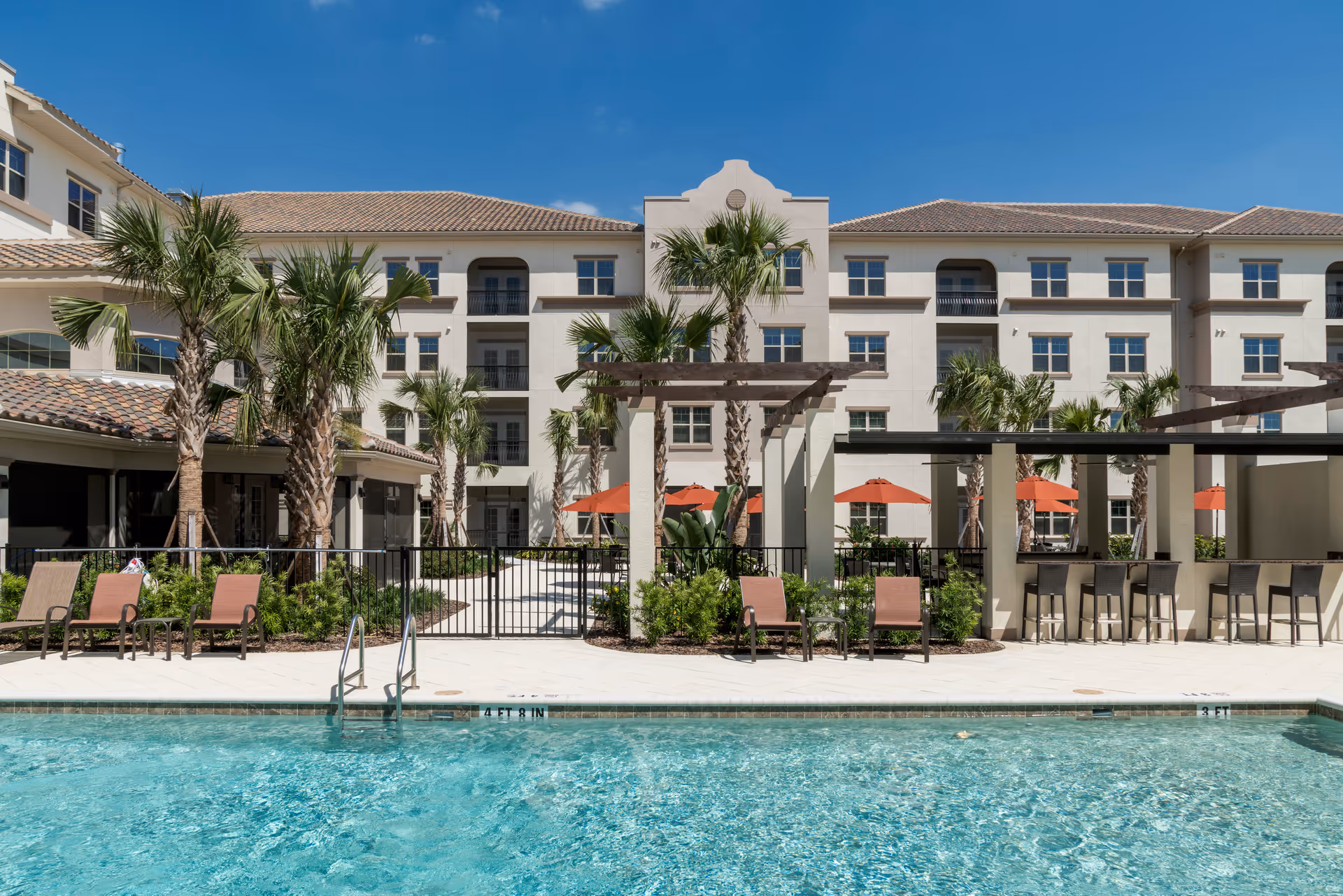 Outdoor swimming pool area with clear blue water, surrounded by lounge chairs and palm trees. In the background, there is a multi-story residential building with balconies and a shaded seating area with orange umbrellas under a pergola.