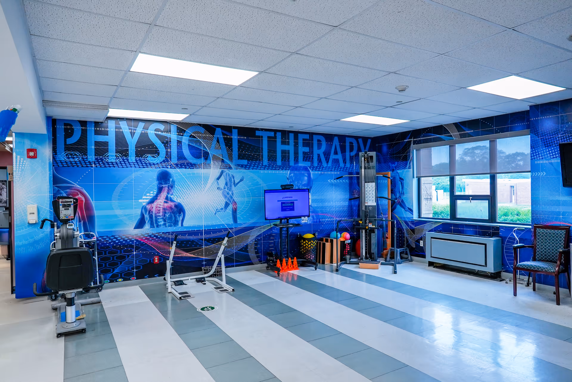 A bright physical therapy room with exercise equipment, cones and therapy tools in front of a large wall mural reading "PHYSICAL THERAPY" and windows on the right.
