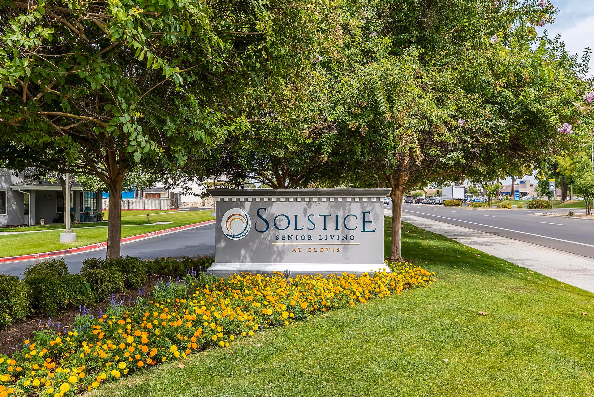 Entrance sign reading "Solstice Senior Living at Clovis" surrounded by trees, flowers, and a lawn next to a street.